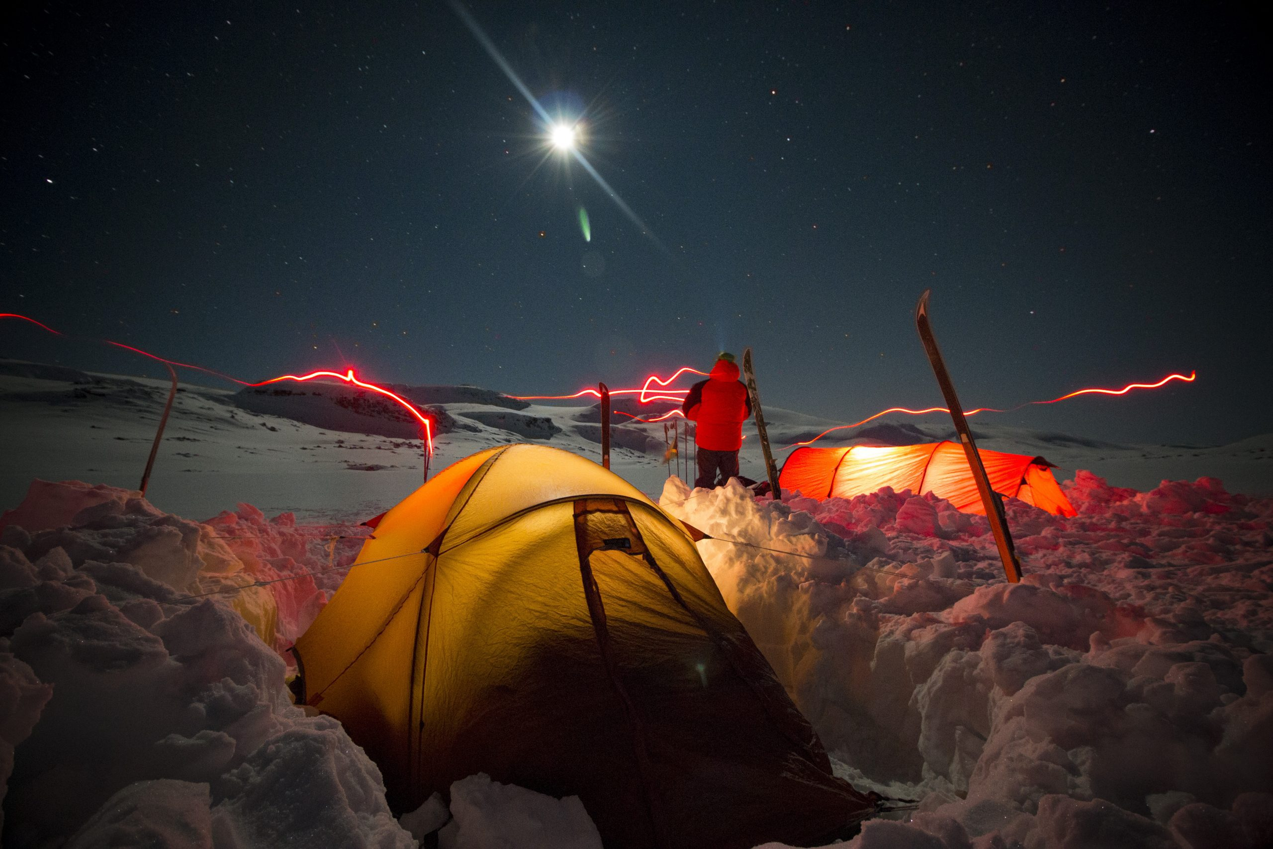 Night-time snowy mountain campsite under a bright moon and starry sky with a glowing yellow tent in the foreground, an illuminated orange tent and a person in a red jacket standing by upright skis amid snow trenches, while red headlamp light trails streak across the scene.