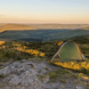 Green one-person tent pitched on a rocky, grassy hilltop at golden hour, overlooking rolling valleys, distant hills and a sunlit reservoir.