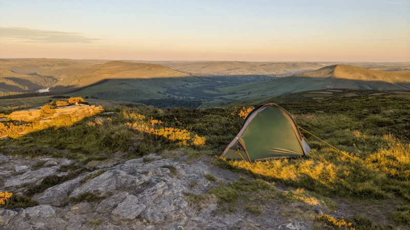 Green one-person tent pitched on a rocky, grassy hilltop at golden hour, overlooking rolling valleys, distant hills and a sunlit reservoir.