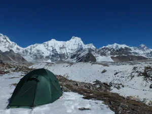 A small green tent pitched on a snowy alpine plateau with jagged, glacier-covered mountains rising under a deep blue sky.