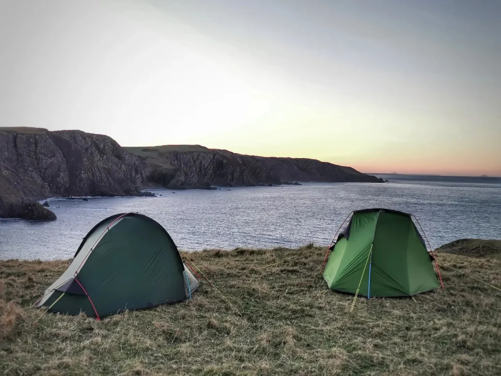 Two green camping tents pitched on a grassy cliff overlooking a calm sea and rugged rocky headlands under a pale sunset sky.