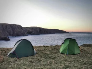 Two green camping tents pitched on a grassy cliff overlooking a calm sea and rugged rocky headlands under a pale sunset sky.