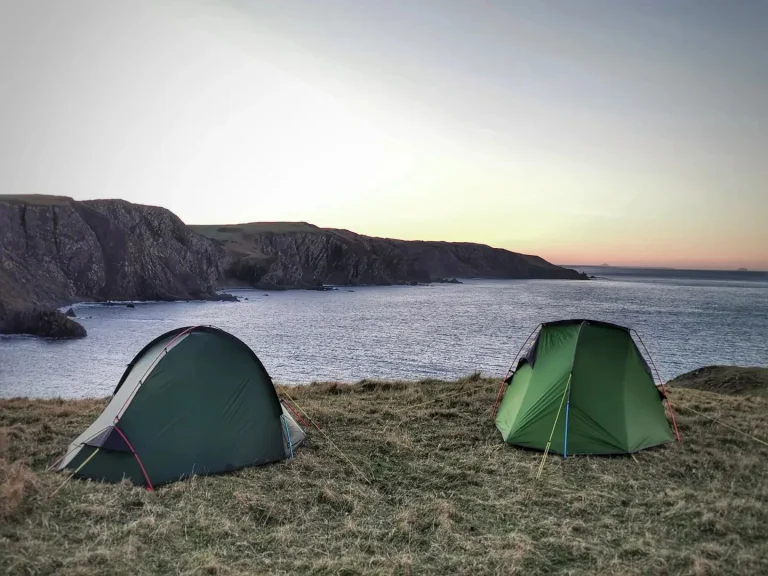 Two green camping tents pitched on a grassy cliff overlooking a calm sea and rugged rocky headlands under a pale sunset sky.