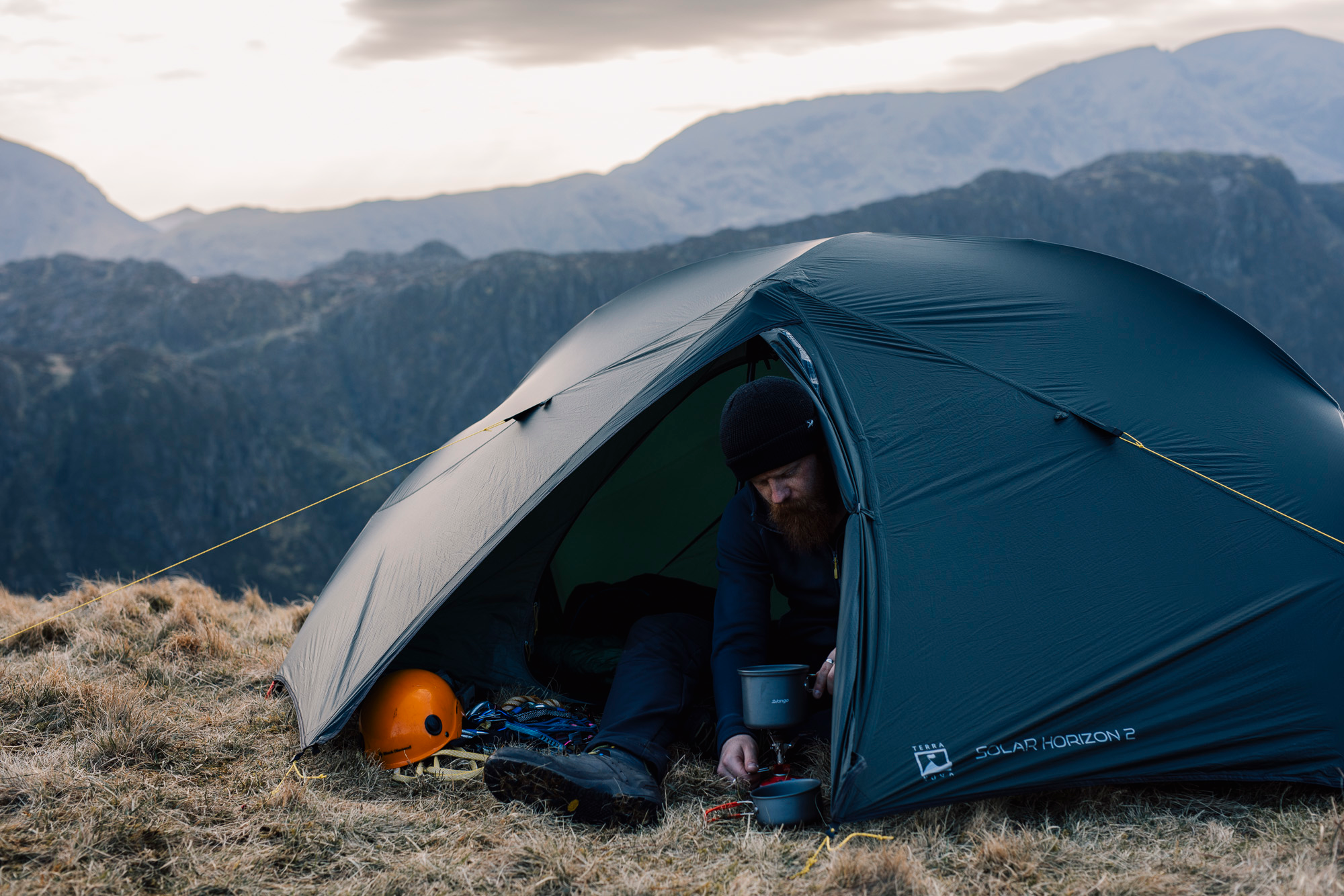 Bearded man in a black beanie seated at the entrance of a dark green tent on a grassy mountain ridge, tending a small camp stove and pots with an orange climbing helmet and ropes beside him and misty layered mountains in the background.