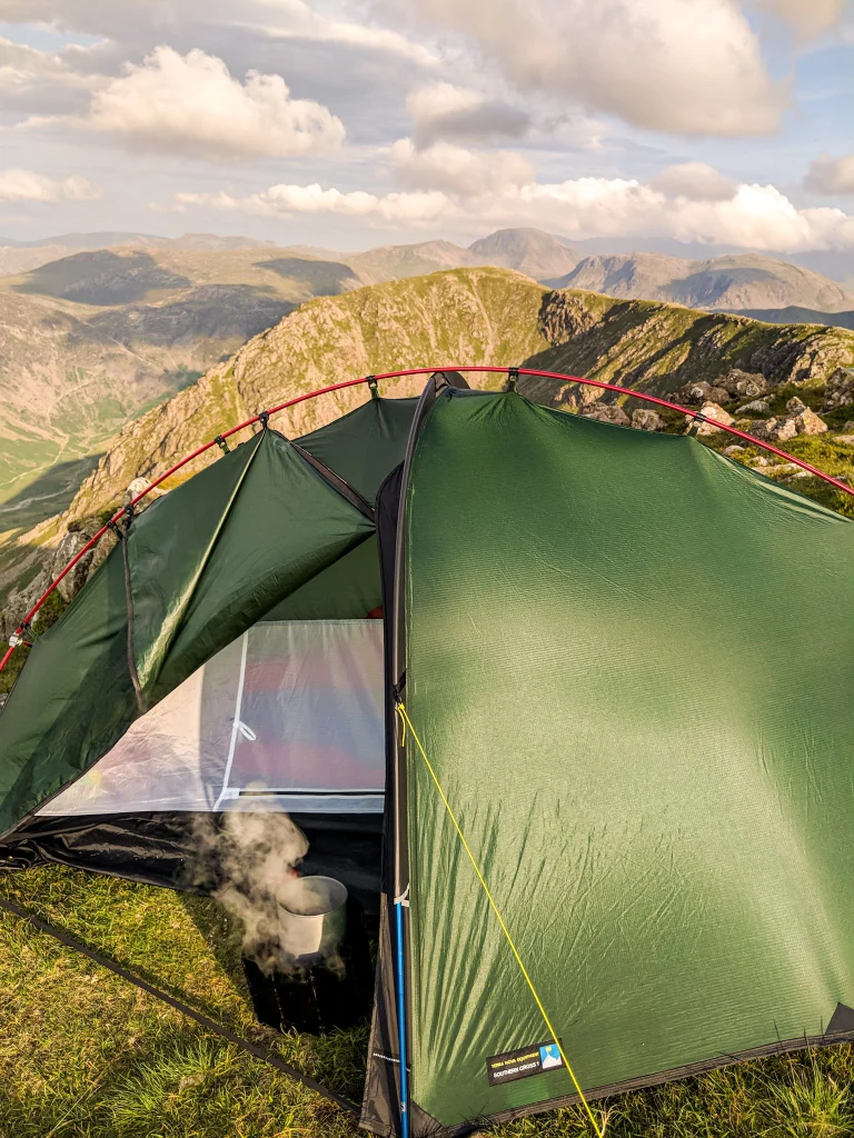 Green backpacking tent pitched on a grassy mountain ridge with red poles and a steaming pot visible inside, overlooking layered rocky peaks under a partly cloudy sky.
