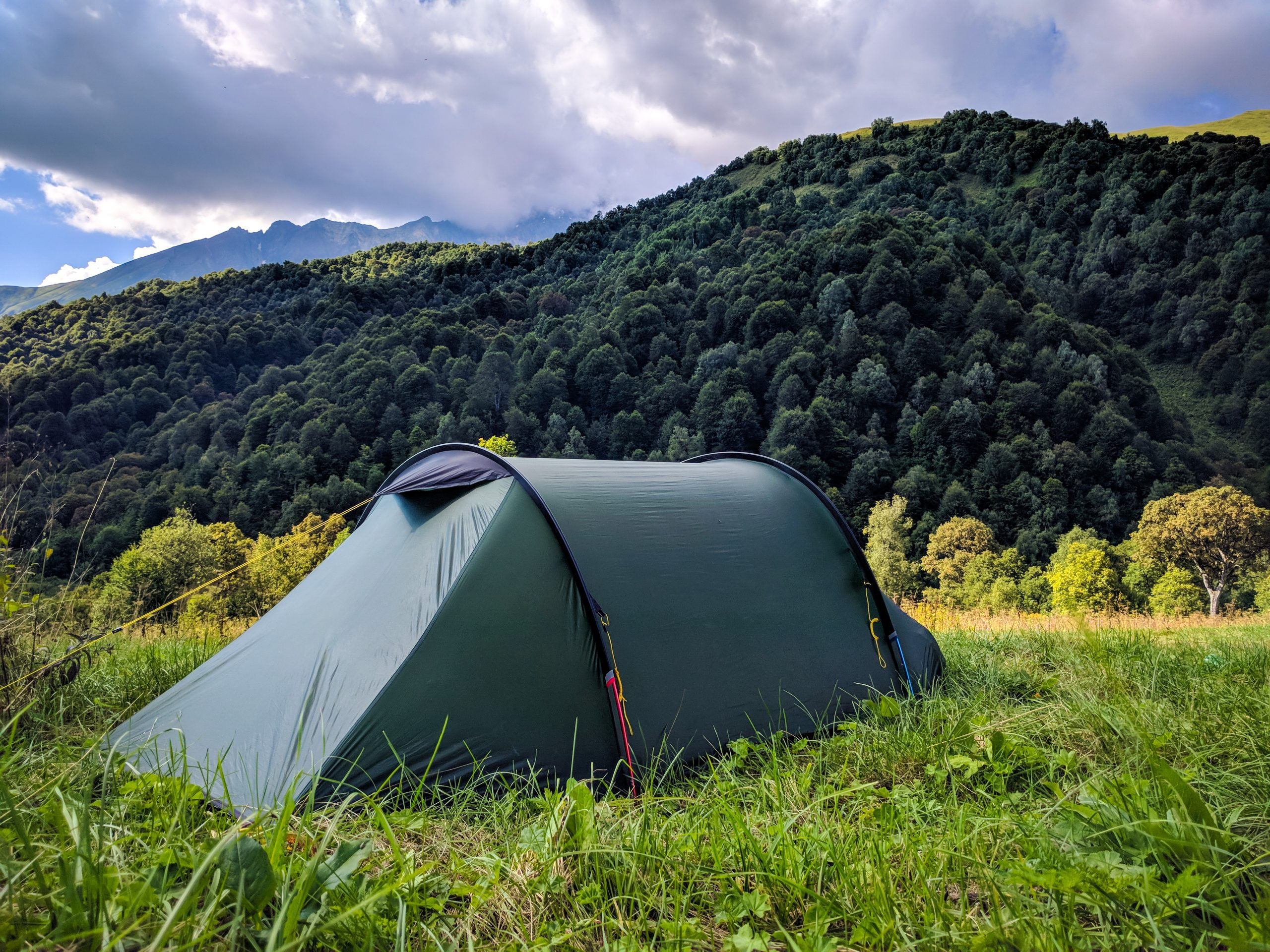 Green camping tent set against a backdrop of lush green mountains and trees under a partly cloudy sky, ideal for outdoor enthusiasts and nature lovers.
