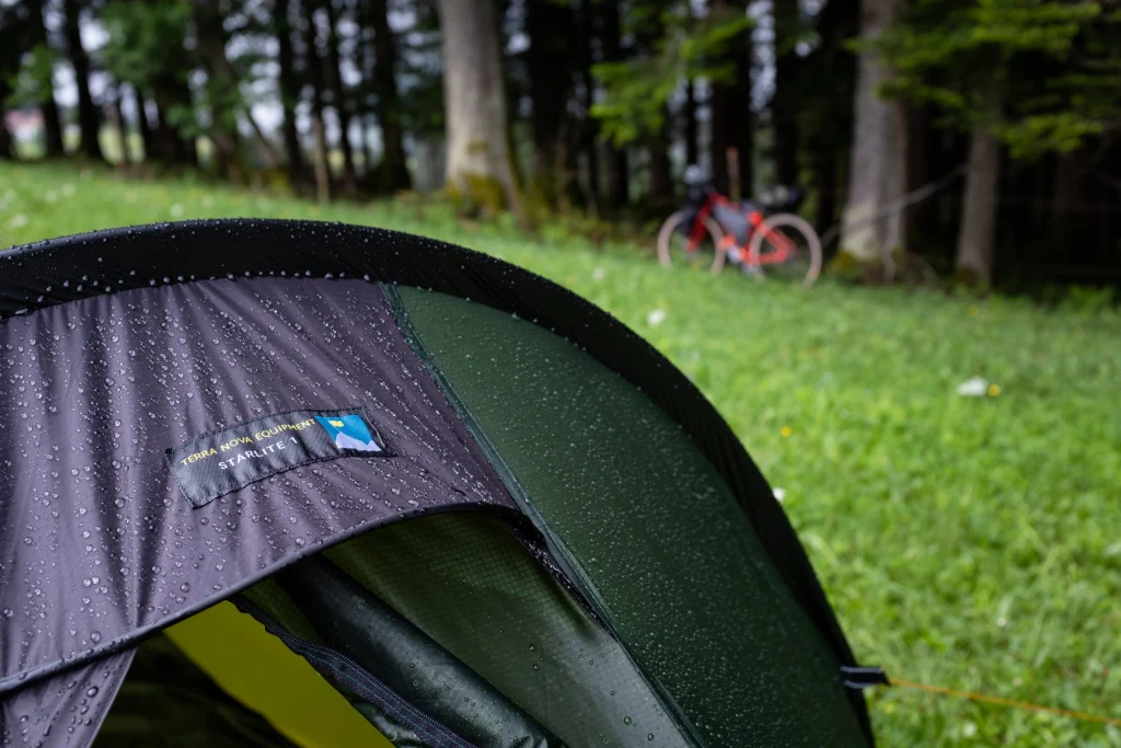 Close-up of a rain-spattered green and grey tent fly showing a Terra Nova Equipment Starlite label, droplets beading on the fabric, with a blurred red bicycle and trees on grassy ground in the background.