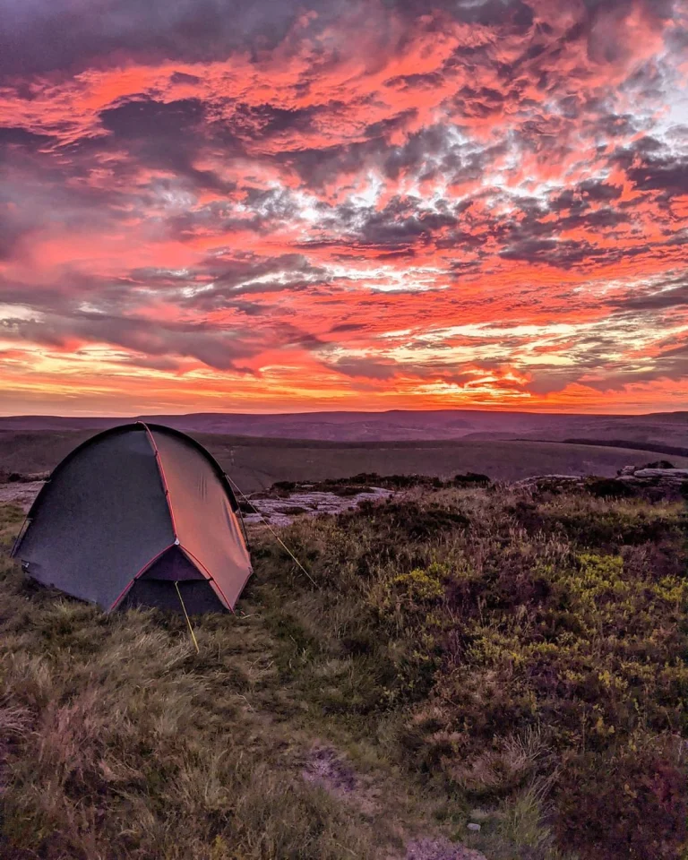 A small dome tent pitched on grassy moorland beneath a dramatic pink and orange sunset sky with layered clouds stretching over distant rolling hills.