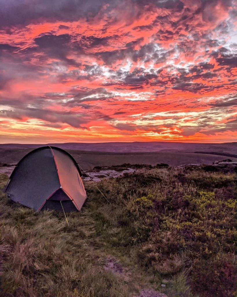 A small dome tent pitched on grassy moorland beneath a dramatic pink and orange sunset sky with layered clouds stretching over distant rolling hills.