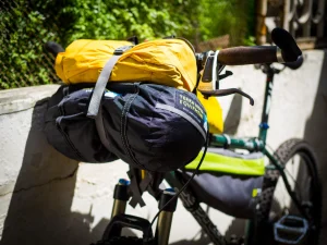 Sunlit bicycle handlebars loaded with a yellow roll-top drybag strapped atop a black Terra Nova Equipment handlebar pack, showing the brake lever, brown grips and part of the green frame and front wheel.