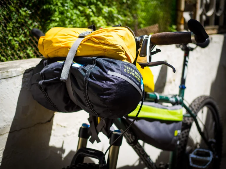 Sunlit bicycle handlebars loaded with a yellow roll-top drybag strapped atop a black Terra Nova Equipment handlebar pack, showing the brake lever, brown grips and part of the green frame and front wheel.