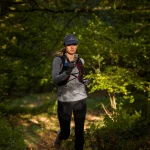 A woman runs along a sun-dappled forest trail wearing a dark cap, grey long-sleeve top, black leggings and a red-trimmed hydration vest, holding a small blue water bottle and wearing gloves.