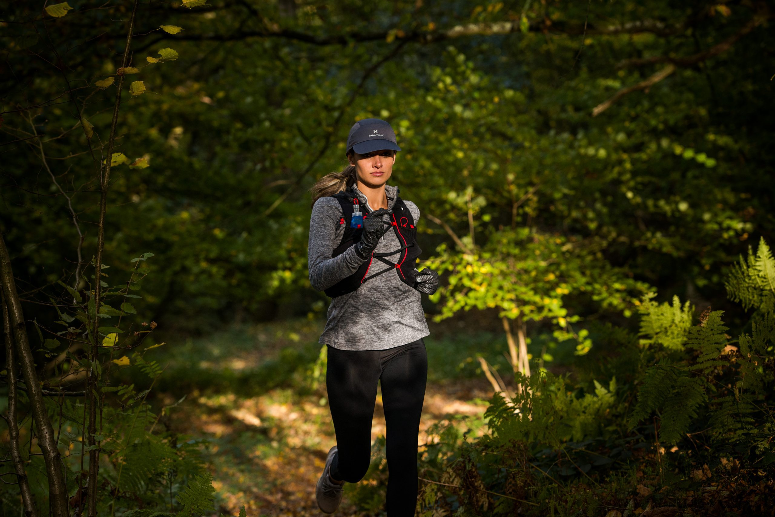 A woman runs along a sun-dappled forest trail wearing a dark cap, grey long-sleeve top, black leggings and a red-trimmed hydration vest, holding a small blue water bottle and wearing gloves.