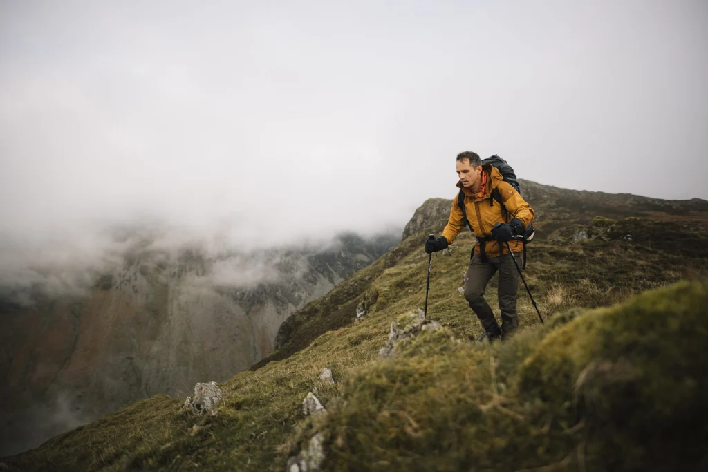 A hiker wearing a yellow jacket and backpack uses trekking poles while ascending a grassy, fog‑shrouded mountain ridge.