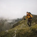 A hiker wearing a yellow jacket and backpack uses trekking poles while ascending a grassy, fog‑shrouded mountain ridge.