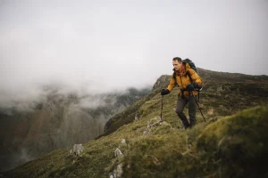 A hiker wearing a yellow jacket and backpack uses trekking poles while ascending a grassy, fog‑shrouded mountain ridge.