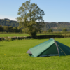 Small green one-person tent pegged with yellow guy lines on a wide grassy field, a lone tree and low dry-stone wall in the midground and tree-covered hills beneath a clear blue sky.