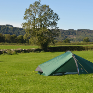 Small green one-person tent pegged with yellow guy lines on a wide grassy field, a lone tree and low dry-stone wall in the midground and tree-covered hills beneath a clear blue sky.
