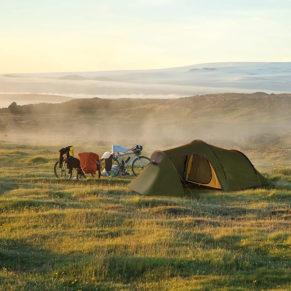 ls_wc_clouds_and_grass-Starlite-3 Green tunnel tent pitched on a misty grassy moor at sunrise beside a touring bicycle with panniers and scattered camping gear, with rolling hills and low fog in the background.