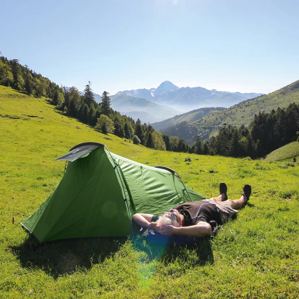 A camper lies on their back beside a small green tent on a sunny grassy mountain meadow, with forested slopes and distant snow-capped peaks under a clear blue sky.