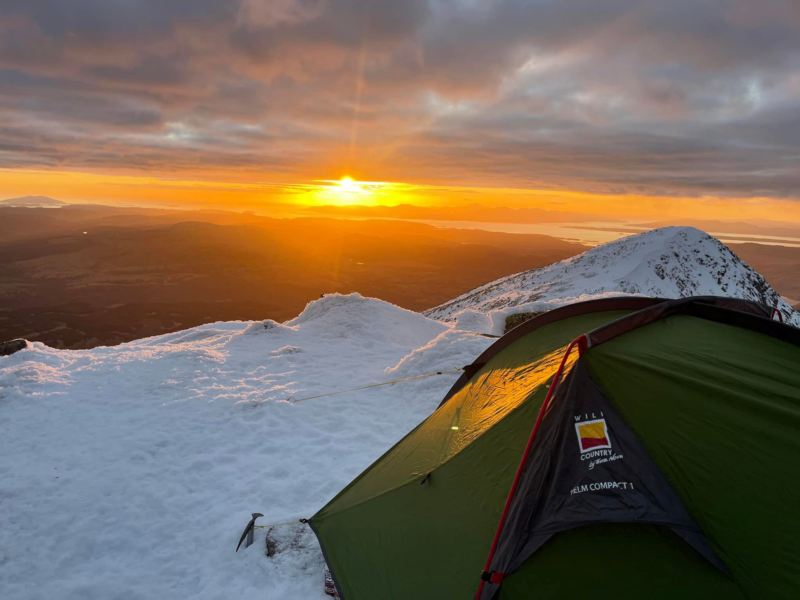 A green backpacking tent staked into snow on a mountain ridge at sunrise, warm orange light spilling across the snowy landscape and distant islands under a cloudy sky with an ice axe planted nearby.
