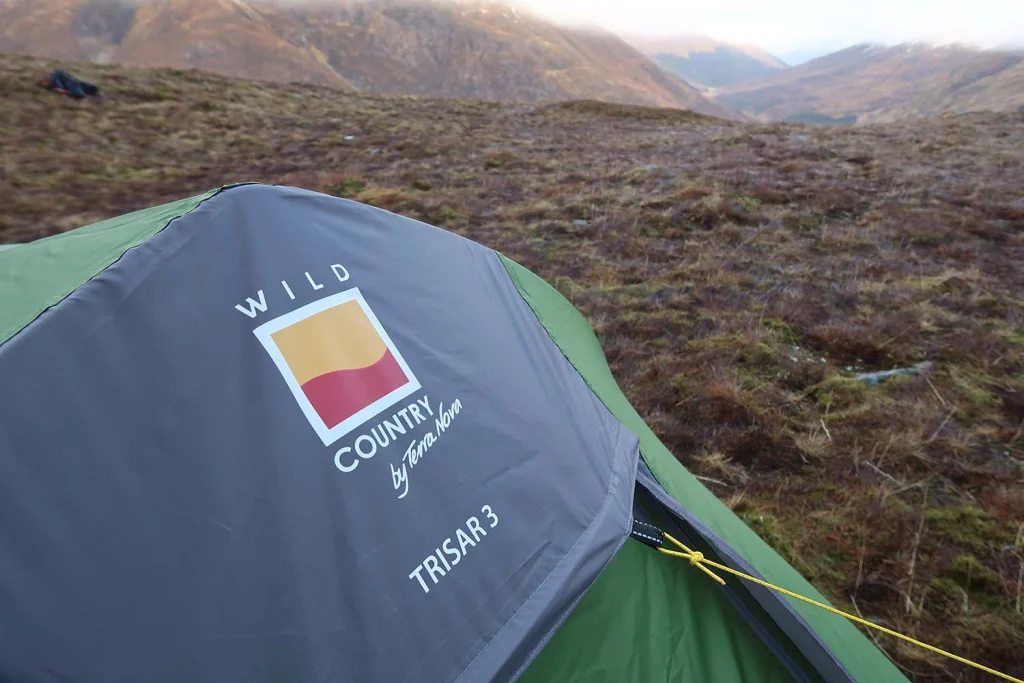 Close-up of a green and grey Wild Country Trisar3 tent fly with the brand logo and a yellow guyline, pitched on brown grassy moorland with rolling hills and a valley visible in the background.