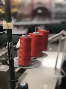 Three large red thread cones on metal spool plates attached to a sewing machine stand, photographed with a shallow depth of field and a blurred textile workshop background