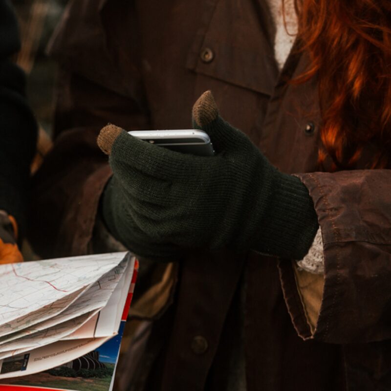 Person wearing green gloves holding a smartphone, with a stack of papers nearby, in a cozy outdoor setting.