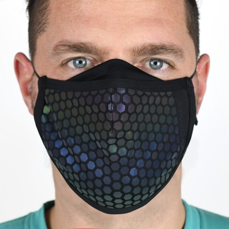 Close-up of a man wearing a black cloth face mask with an iridescent hexagonal honeycomb pattern, showing his light blue eyes and short dark hair against a plain background.
