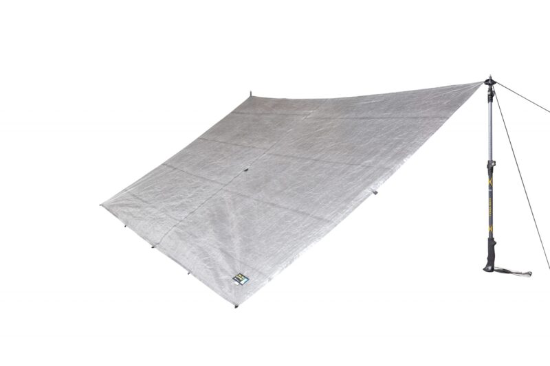 Rectangular silver camping tarp pitched at an angle and anchored by a trekking pole and guyline, showing reinforced corner tabs and a small brand label on a plain white background.