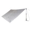 Silver rectangular tarp shelter with reinforced attachment points and a small brand label, pegged at three corners and tensioned on the right by a walking pole and guy line.