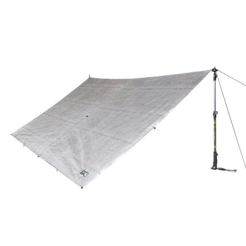 Silver rectangular tarp shelter with reinforced attachment points and a small brand label, pegged at three corners and tensioned on the right by a walking pole and guy line.