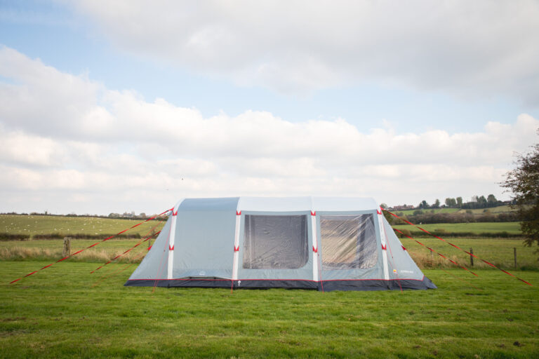 Large light-grey family tent with red guy ropes and clear plastic windows pitched on a grassy field with rolling hills and a cloudy sky in the background.