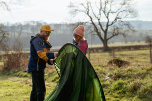 Couple setting up a green tent outdoors in a rural area, surrounded by trees and fields, on a sunny day.