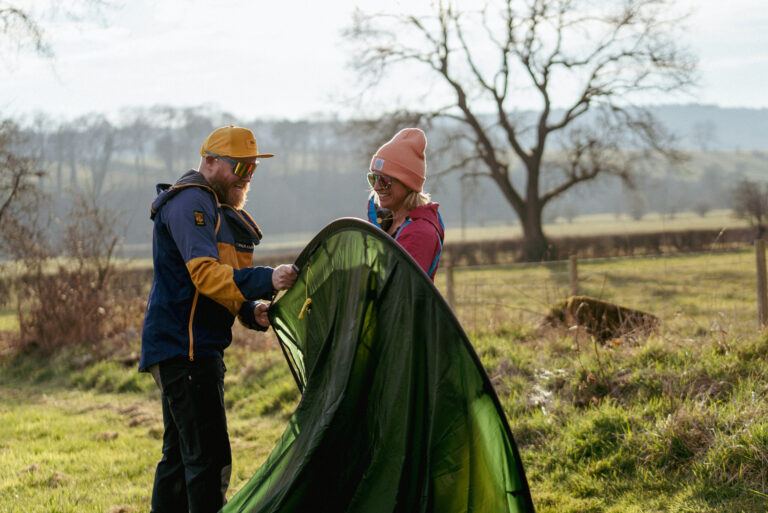 Couple setting up a green tent outdoors in a rural area, surrounded by trees and fields, on a sunny day.