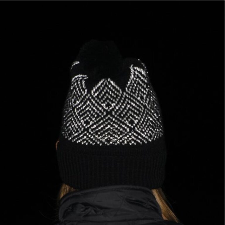 Rear view of a person wearing a black knitted pompom hat with a white reflective zigzag pattern and a turned-up ribbed brim, black jacket collar and a few strands of hair visible against a dark background.