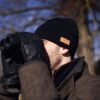 Man in a black beanie and leather gloves holding binoculars to his eyes while wearing a brown winter coat outdoors against a blurred background of bare trees.