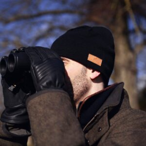 Man in a black beanie and leather gloves holding binoculars to his eyes while wearing a brown winter coat outdoors against a blurred background of bare trees.