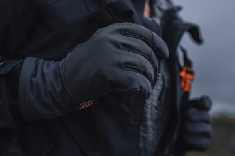 Close-up of a person's hand wearing a black outdoor glove, showcasing its stylish design and functionality for cold weather activities. The glove is part of a weather-resistant jacket ensemble, ideal for hiking or winter sports.