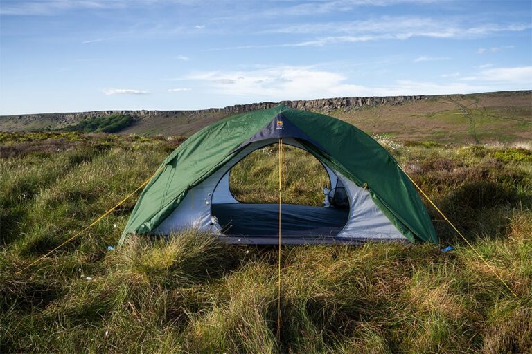 Green dome tent with its entrance open, staked and yellow guy‑lined on windswept grassy moorland with a low rocky escarpment and blue sky in the background.