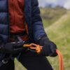 Close-up of a person in a navy puffer jacket and black gloves threading an orange climbing rope through an orange belay device and carabiner while clipped into a harness on a grassy hillside