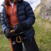 Person wearing a navy puffer jacket and orange zip-up adjusts an orange climbing rope and gold carabiners with black gloves on a grassy rocky hillside.
