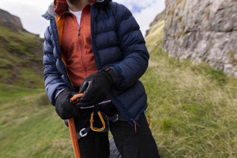 Person wearing a navy puffer jacket and orange zip-up adjusts an orange climbing rope and gold carabiners with black gloves on a grassy rocky hillside.
