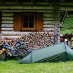 Green one-person tent pitched on grass beside a rustic log cabin with stacked firewood; a fully loaded touring bicycle leans against the cabin and a wooden picnic table with helmets and gear sits nearby, with a grassy forest slope in the background.