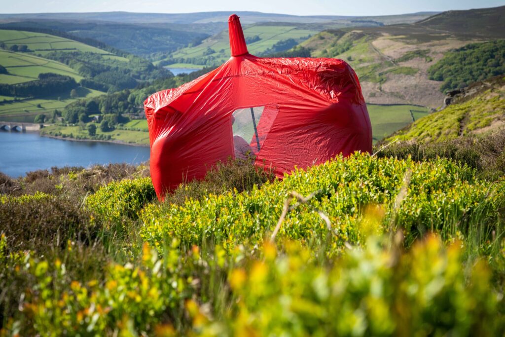 A bright red tent wrapped in a taut protective cover sits among green heather on a sunlit moorland slope, overlooking a blue reservoir and patchwork rolling hills.