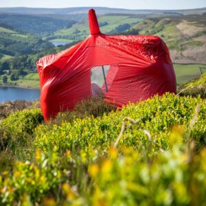 A bright red tent wrapped in a taut protective cover sits among green heather on a sunlit moorland slope, overlooking a blue reservoir and patchwork rolling hills.
