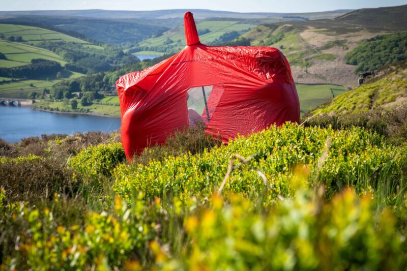 A bright red tent wrapped in a taut protective cover sits among green heather on a sunlit moorland slope, overlooking a blue reservoir and patchwork rolling hills.
