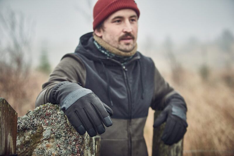A man wearing a red beanie, scarf and dark jacket leans on a lichen-covered stone gatepost in a misty field, his black-gloved hand prominent in the foreground.