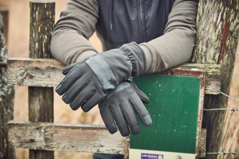A pair of dark grey insulated gloves on crossed hands resting over a weathered wooden fence next to a green sign, the wearer's grey jacket sleeves visible.