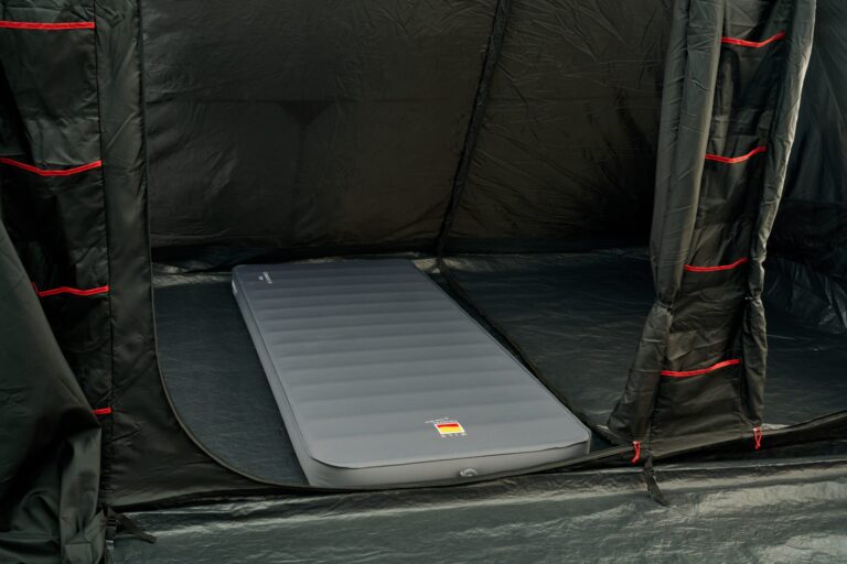Interior of a black camping tent with a single grey inflatable sleeping mat laid on the floor and black fabric walls featuring vertical red‑trimmed storage pockets.